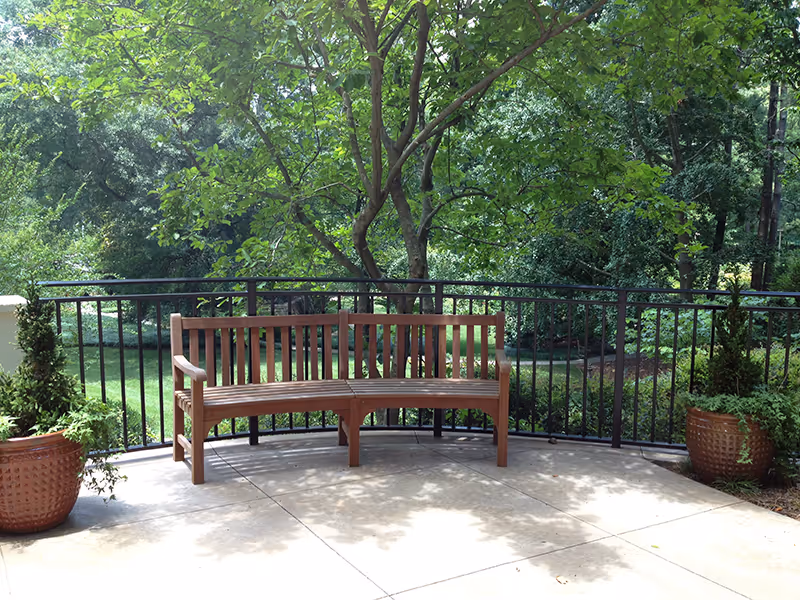 A curved wooden bench on a concrete patio surrounded by a black metal railing. There are two large potted plants on either side of the bench and lush green trees and grass in the background.