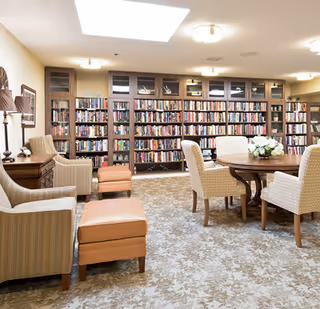 Cozy common room with bookshelves along the back wall, a round table with chairs, and upholstered armchairs with ottomans.