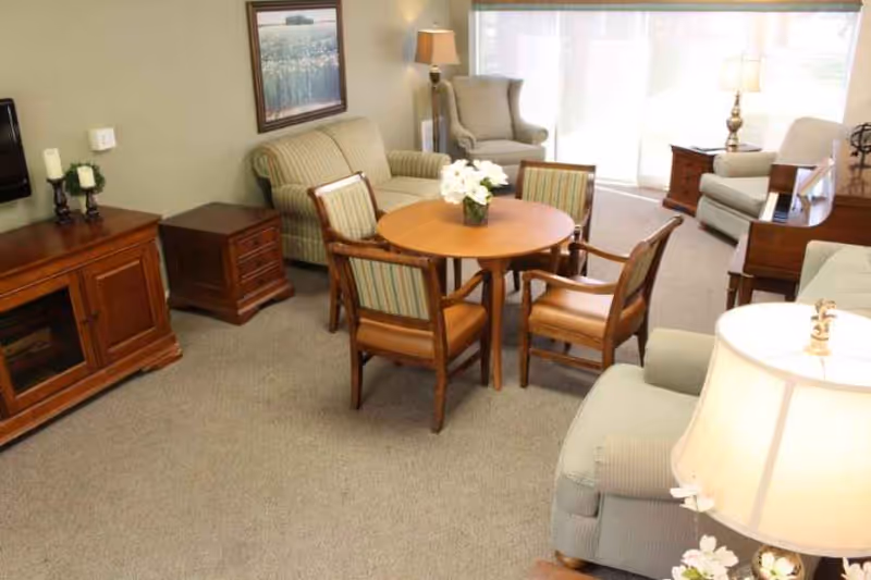 A cozy living room area with a round wooden table surrounded by four cushioned chairs. The room features multiple upholstered armchairs and a sofa, a wooden TV stand with a television, side tables with lamps, and a large window with blinds allowing natural light to fill the space. A framed picture hangs on the wall above the sofa.