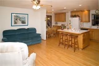 Interior view of a living space featuring a green couch, a white armchair, a kitchen island with two wooden stools, wooden kitchen cabinets, a dining table with chairs, and a ceiling fan with lights.