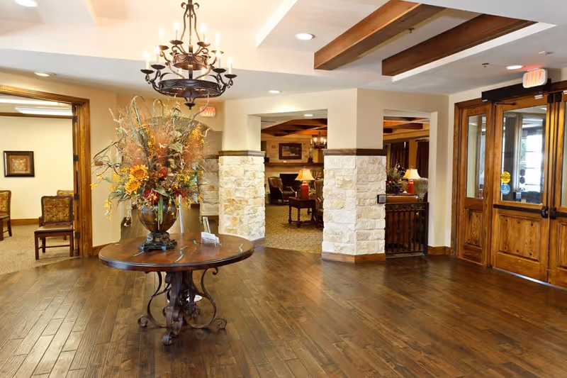 A spacious and warmly lit interior area with wooden flooring and ceiling beams. In the center, there is a round wooden table with an ornate metal base holding a large floral arrangement. To the right, there are wooden double doors with glass panels leading outside. The background shows a cozy seating area with lamps and stone pillars partially dividing the space.
