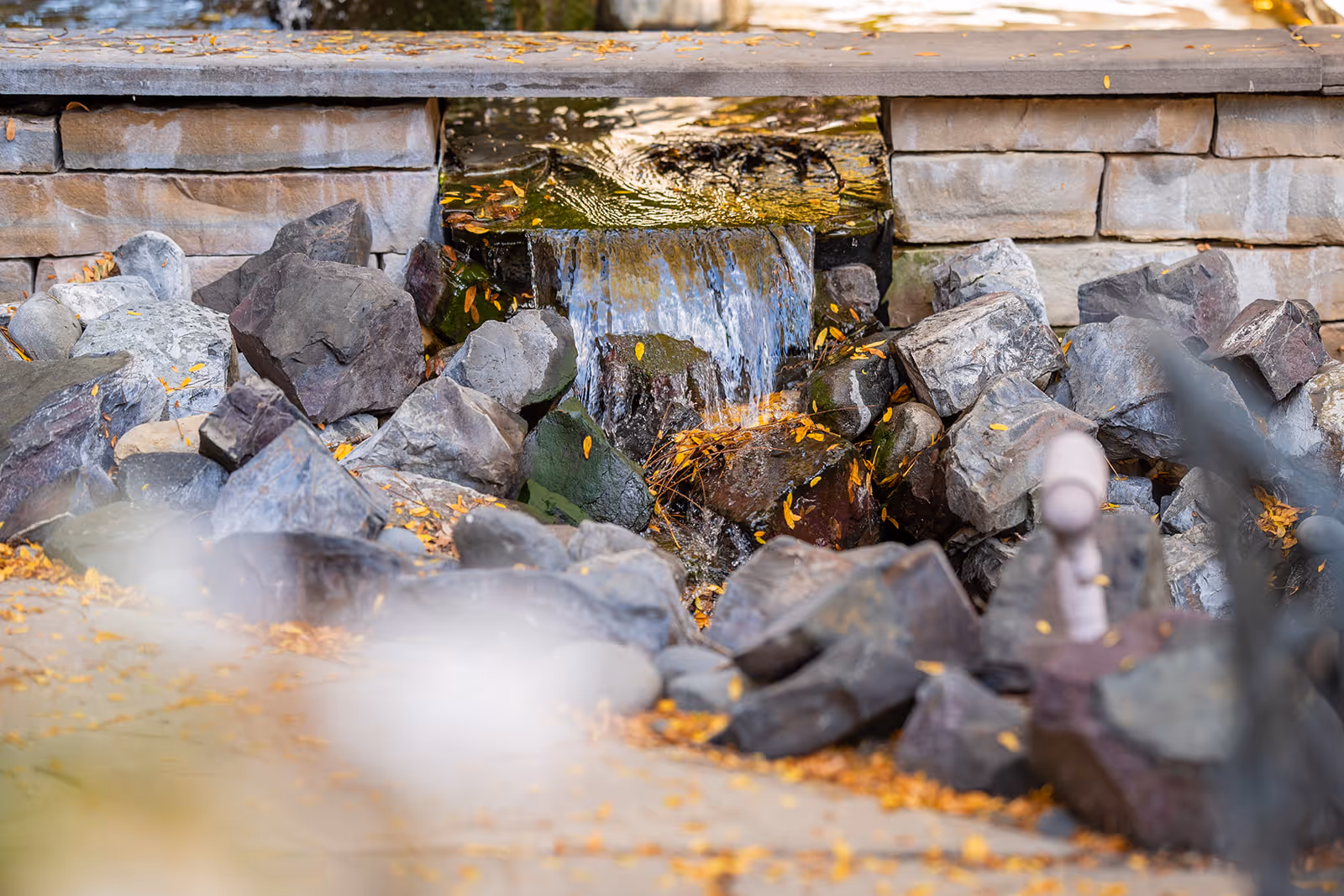 Small outdoor stone waterfall feature with water flowing over a ledge into rocks below, scattered with fallen leaves.