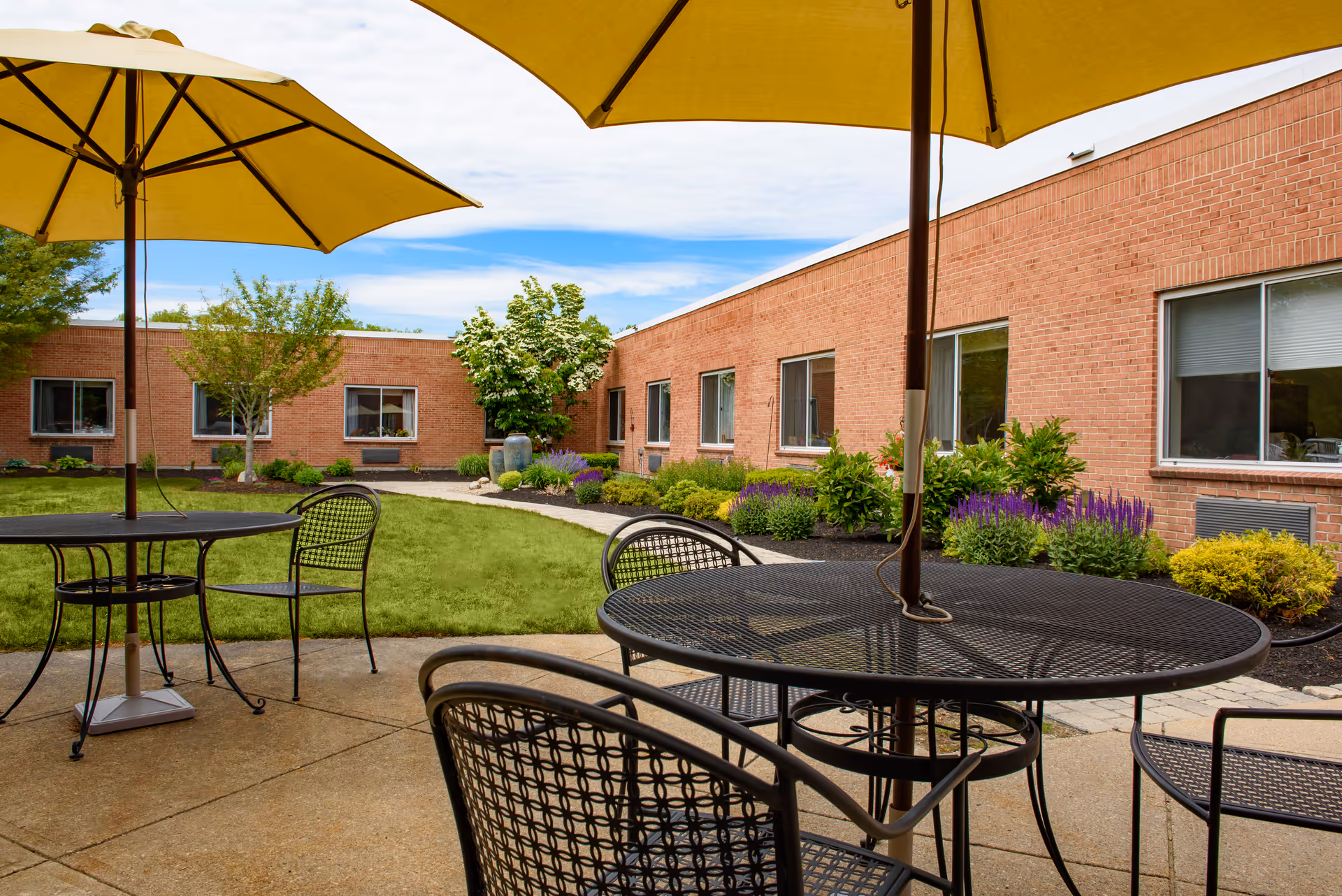 Outdoor patio area with black metal tables and chairs under large yellow umbrellas. The patio is adjacent to a single-story brick building with several windows. There is a well-maintained lawn and landscaped garden beds with shrubs and flowers surrounding the patio area.