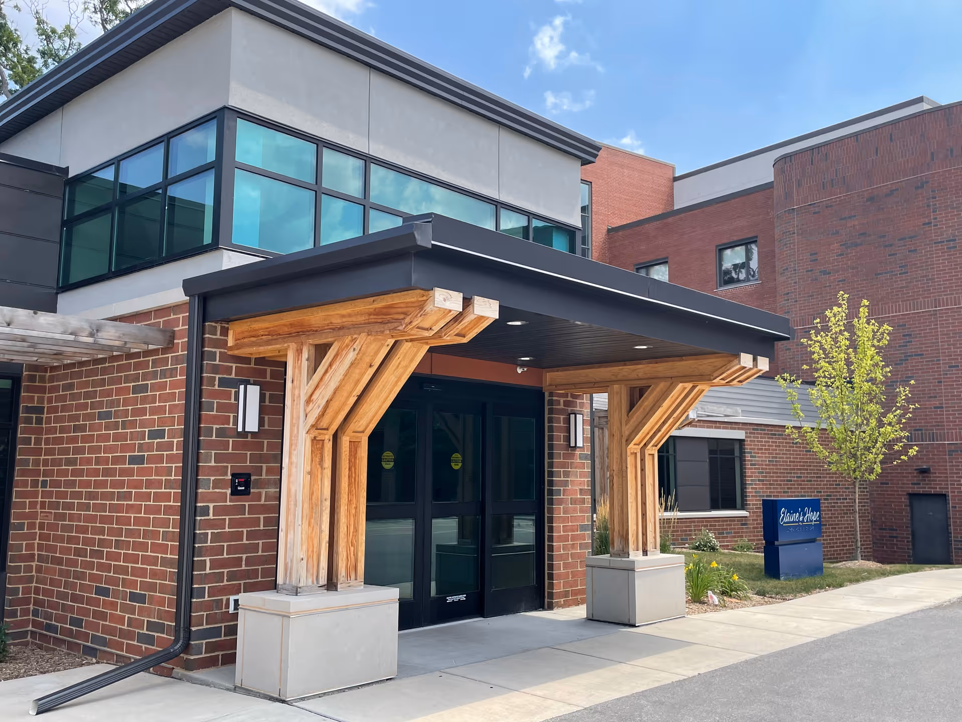 Front entrance of a brick building with a wooden-pillared canopy over glass double doors and a small blue sign to the right.