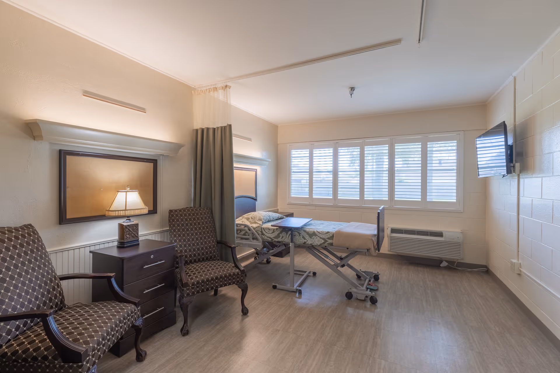 A bright and clean patient room with a hospital bed, two patterned armchairs, a small bedside table with a lamp, a wall-mounted TV, and large windows with white shutters letting in natural light.