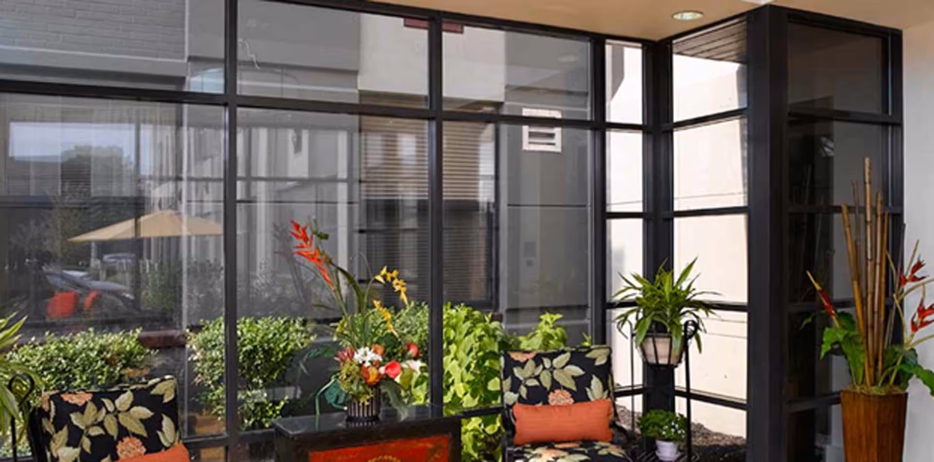 Sunlit indoor seating area with floral-upholstered chairs, potted plants, and large floor-to-ceiling windows.