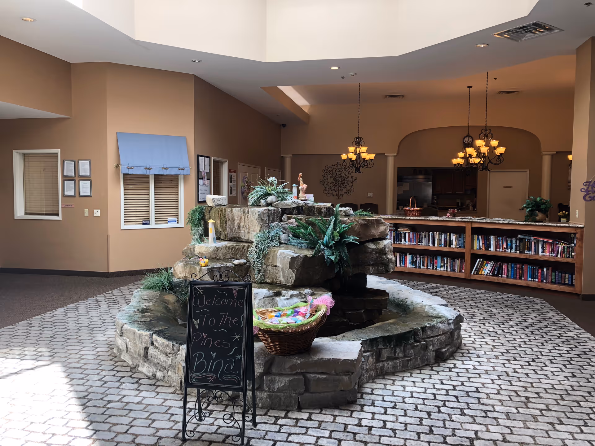 Interior view of a senior living facility lobby area featuring a stone water fountain decorated with plants and a basket of colorful items. A blackboard sign in front of the fountain reads 'Welcome to The Pines Bingo.' Behind the fountain is a bookshelf filled with books and a kitchen area with hanging chandeliers. The walls are painted beige and the floor has a cobblestone pattern.