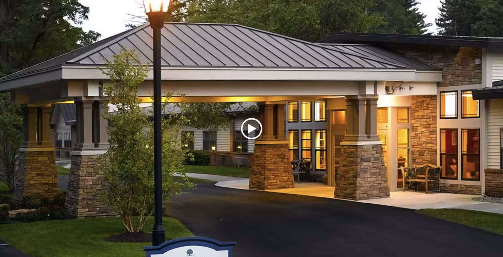 Exterior view of Peregrine Senior Living at Guilderland showing a covered entrance with stone pillars and a metal roof, illuminated by warm lights in the evening. There is a lamppost and some greenery around the driveway leading to the entrance.