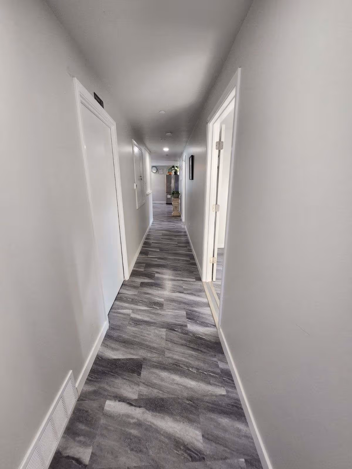 A long, narrow hallway with gray and white marbled flooring and white walls. There are several white doors along the hallway, and at the far end, a cabinet with plants and a clock on the wall is visible. The hallway is well-lit with ceiling lights.