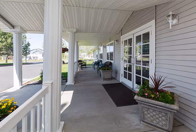 Covered outdoor porch area with white columns, gray siding walls, potted plants, and several chairs along the walkway. The porch overlooks a green lawn and a paved road.