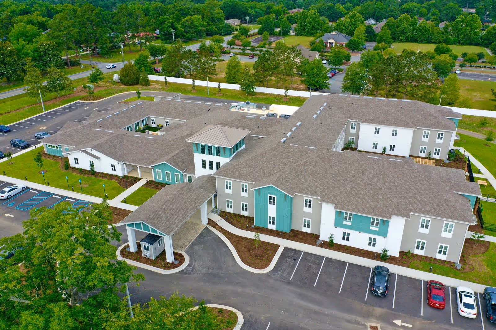 Aerial view of The Canopy at Azalea Grove, a large senior living facility with multiple connected buildings featuring gray roofs and white and teal exterior walls. The facility is surrounded by parking lots, green lawns, trees, and nearby roads.