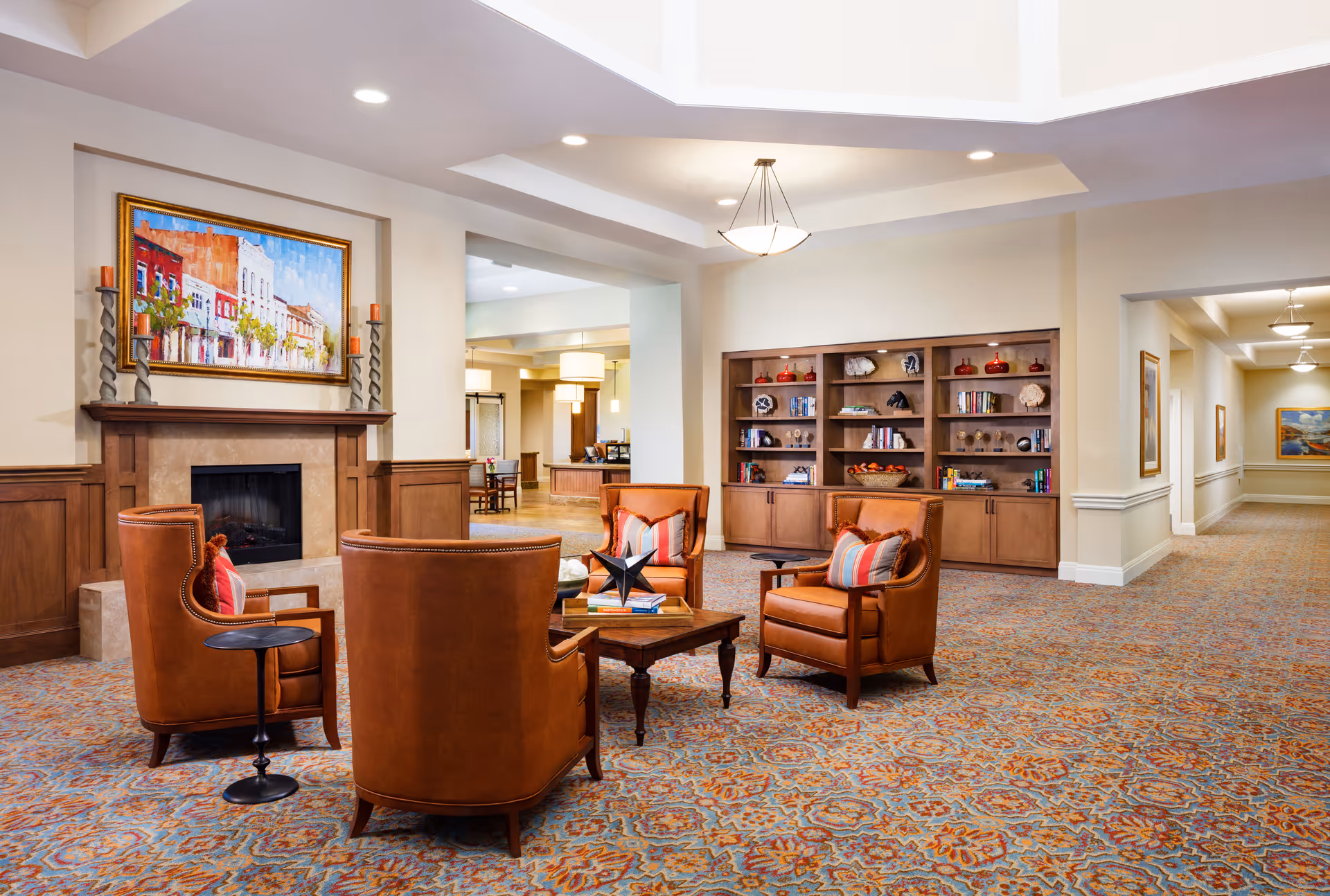 A spacious and well-lit common area with four brown leather armchairs arranged around a wooden coffee table. The room features a fireplace with a colorful painting above it, built-in wooden bookshelves filled with books and decorative items, and patterned carpet flooring. The ceiling has recessed lighting and a hanging light fixture, with hallways visible in the background.