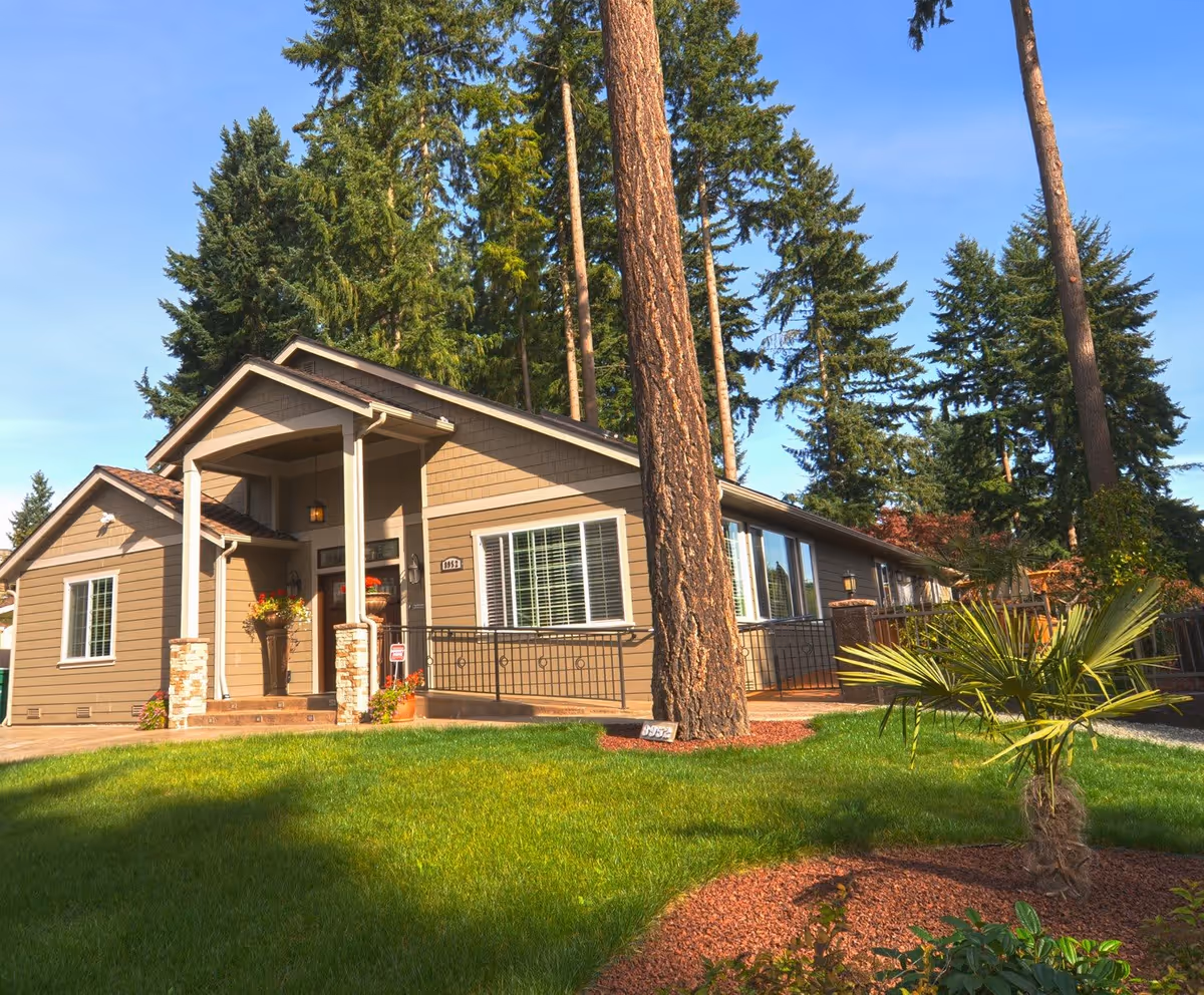 Exterior view of a single-story residential building with beige siding and white trim, surrounded by tall trees and a well-maintained green lawn with landscaped plants. The entrance features a small porch with two columns and potted plants.