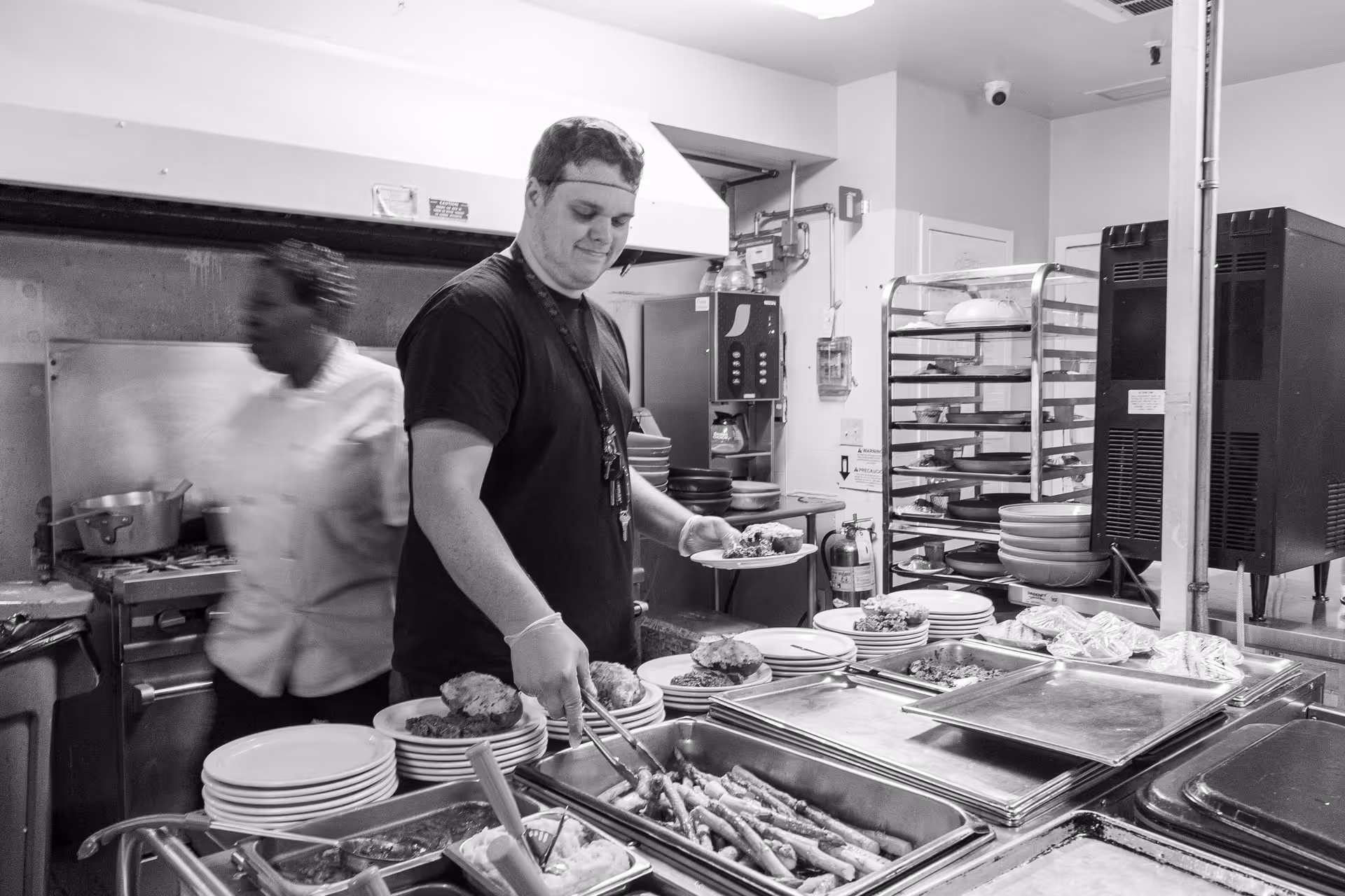 A man serving food onto a plate in a kitchen setting, with a woman in a chef's coat moving in the background. The kitchen has trays of food, stacks of plates, and kitchen equipment visible.