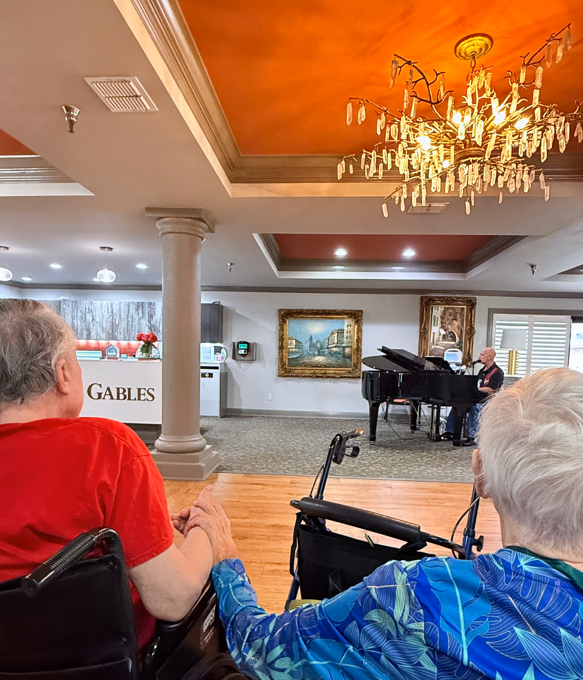 Two elderly individuals sitting in wheelchairs holding hands in a spacious, well-lit room with a grand piano and a pianist performing. The room features an orange ceiling with a decorative chandelier, framed paintings on the walls, and a reception desk labeled 'GABLES'.