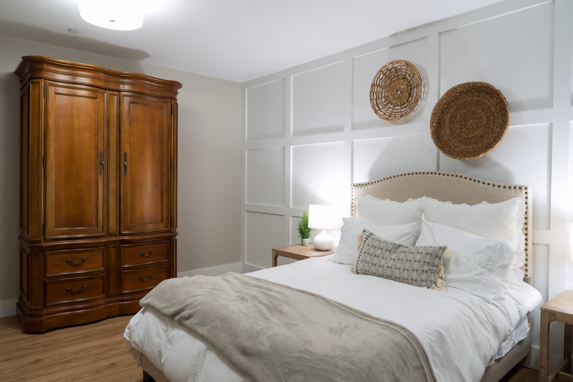 A cozy bedroom with a large wooden armoire on the left and a neatly made bed with white linens and multiple pillows. The bed has a beige upholstered headboard with nailhead trim. Two woven decorative baskets hang on the white paneled wall above the bed. There are wooden nightstands on each side of the bed, each with a small lamp and a plant on the left nightstand.
