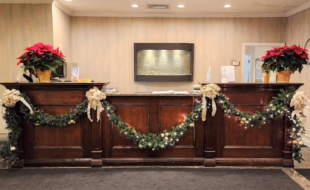 Reception desk decorated with Christmas garlands, white bows, and poinsettia plants on each end. Behind the desk is a wall with a mounted water feature and a doorway to the right.