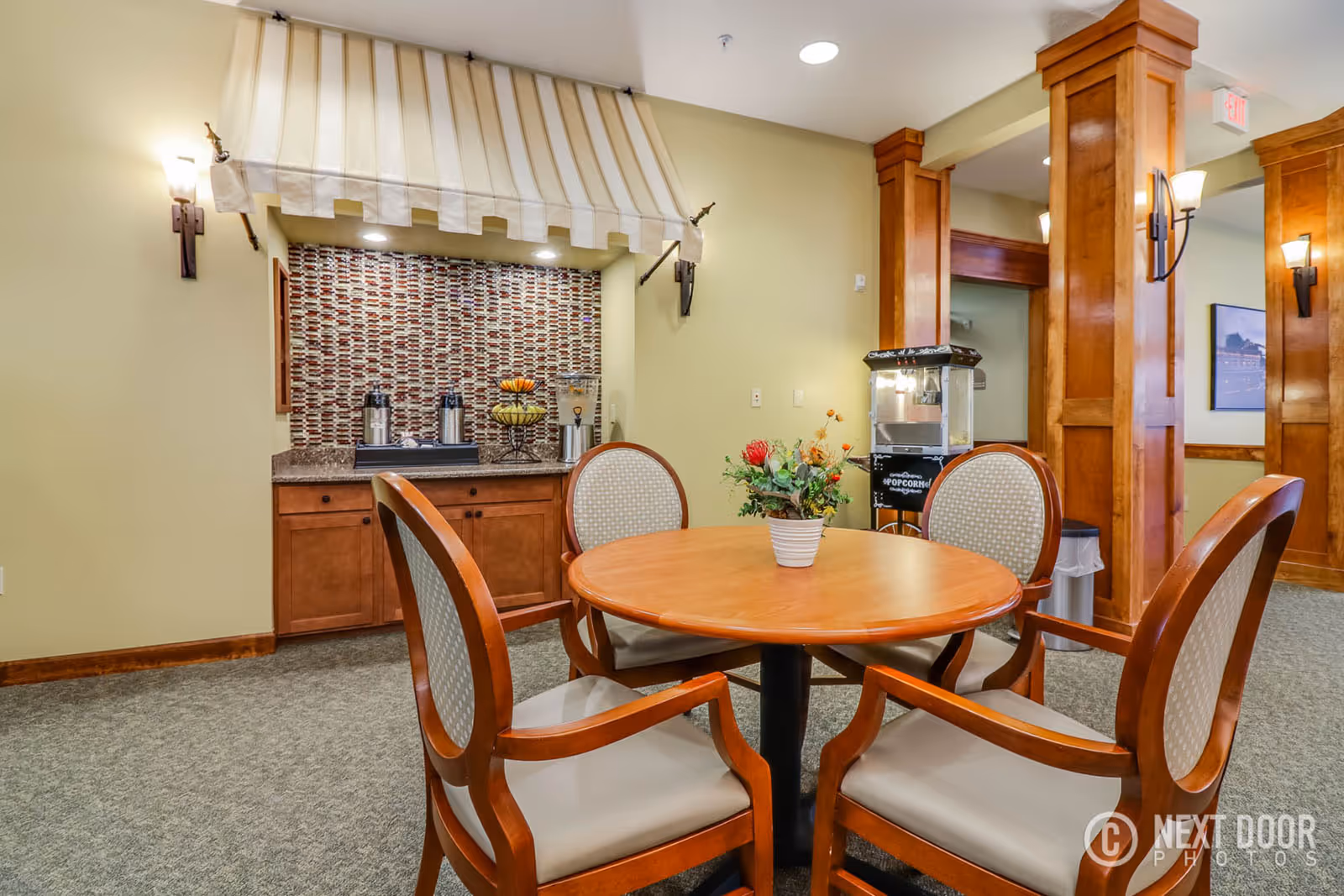 A cozy seating area in an assisted living facility with a round wooden table surrounded by four cushioned chairs. On the table is a small floral arrangement. Behind the table is a counter with a mosaic tile backsplash, two beverage dispensers, a fruit stand, and a water dispenser. To the right, there is a popcorn machine next to wooden pillars and wall sconces providing warm lighting.