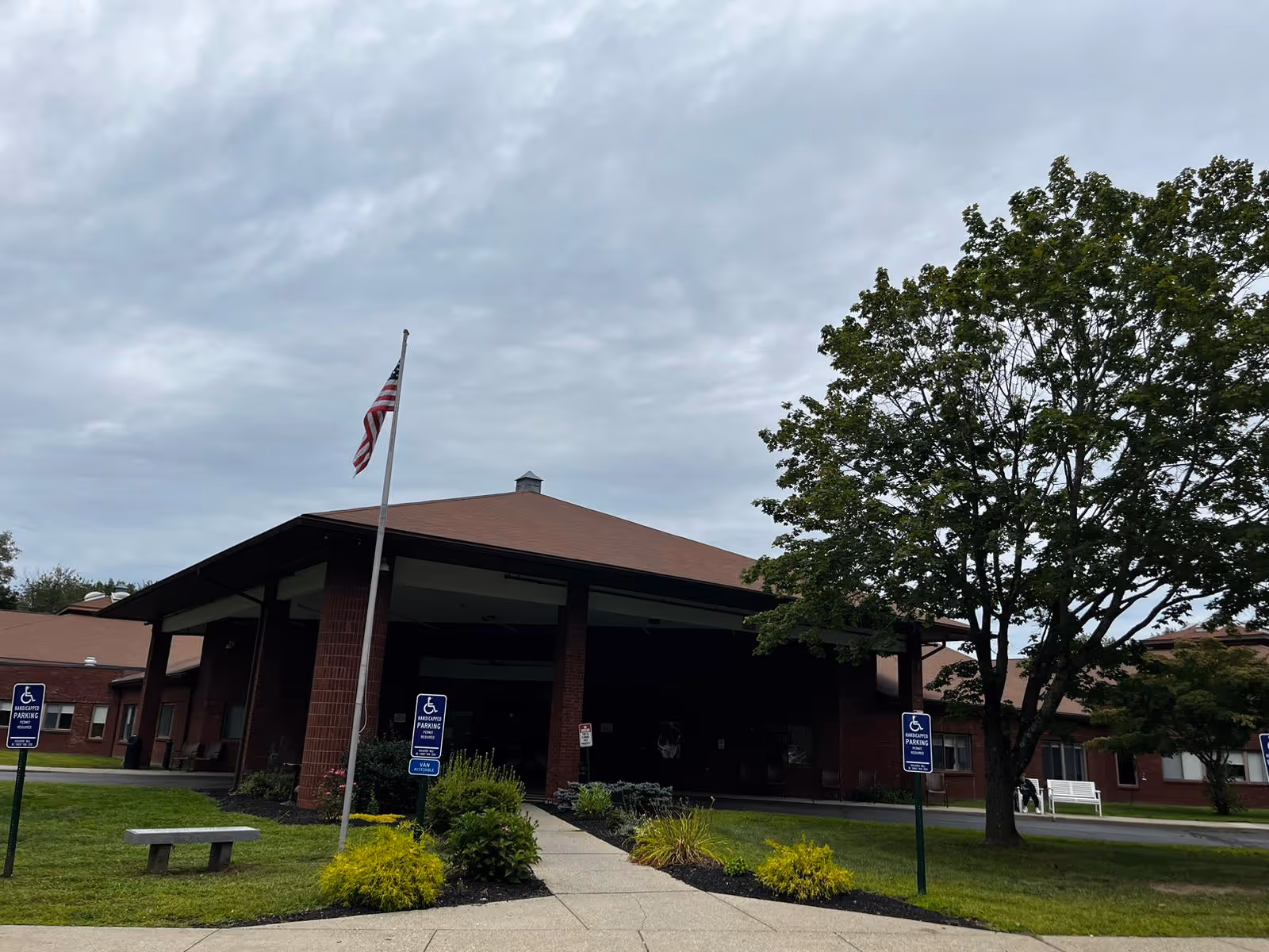 Front entrance of a brick senior living building with a covered drive, flagpole, accessible parking signs, and a large tree.
