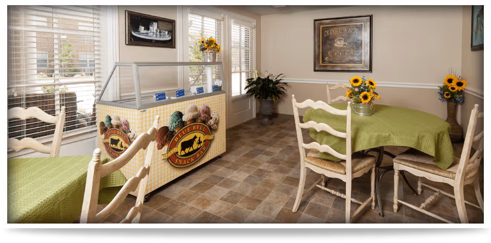 A cozy dining area with two tables covered in green tablecloths and surrounded by wooden chairs. There is a Blue Bell snack bar freezer against the wall, decorated with images of ice cream cones. The room has large windows with white blinds, potted plants, and framed artwork on the walls. The floor is tiled with a brown and beige pattern.