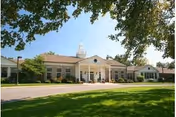 Front exterior of a single-story senior living facility with a columned entrance, cupola, and manicured lawn under trees.