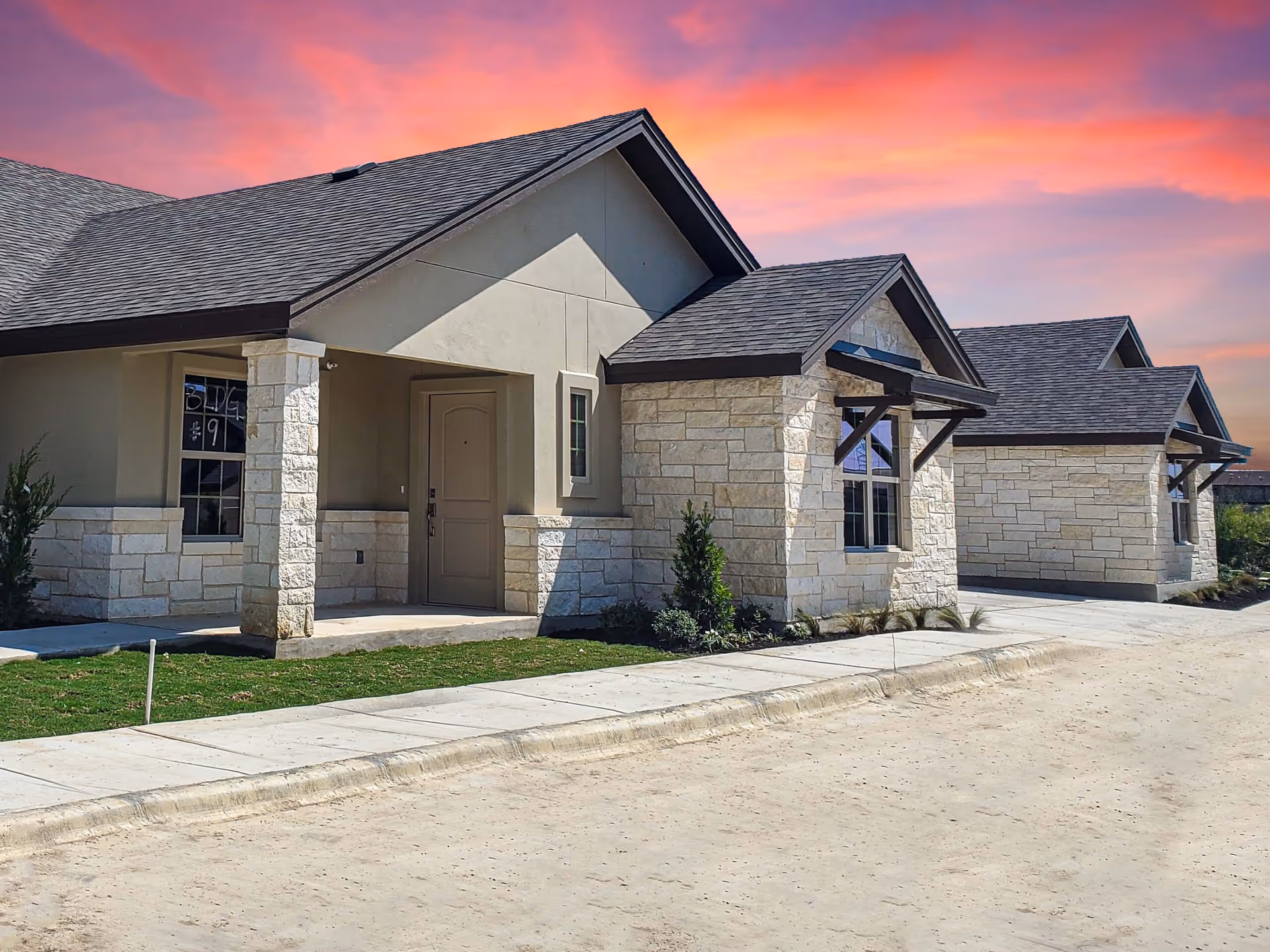 Exterior view of single-story residential cottages with stone and stucco walls, dark shingled roofs, and small landscaped areas under a vibrant pink and orange sunset sky.