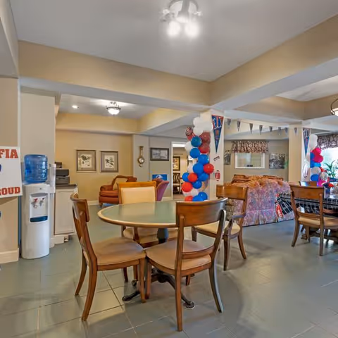A communal living area in a senior living facility decorated with red, white, and blue balloons and pennants. The room features round wooden tables with chairs, a water cooler, framed pictures on the walls, and comfortable seating with sofas in the background. The space is well-lit with ceiling lights and has a tiled floor.