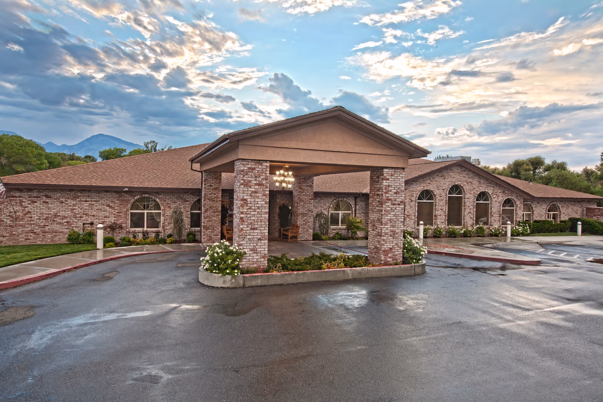 Front entrance of a brick senior living facility with a covered porte-cochère, arched windows, and a wet driveway under a cloudy sky.