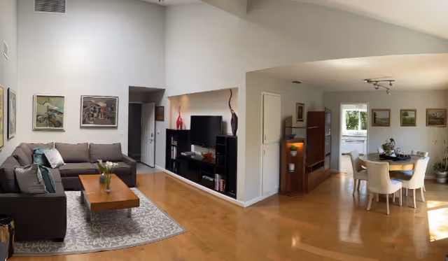 A spacious and well-lit living and dining area with wooden floors. The living room features a gray sectional sofa with cushions, a wooden coffee table on a patterned rug, and a TV on a black entertainment unit. The dining area has a round table with six white upholstered chairs and is decorated with framed paintings on the wall. There is a doorway leading to a kitchen area with white cabinets and a window letting in natural light.