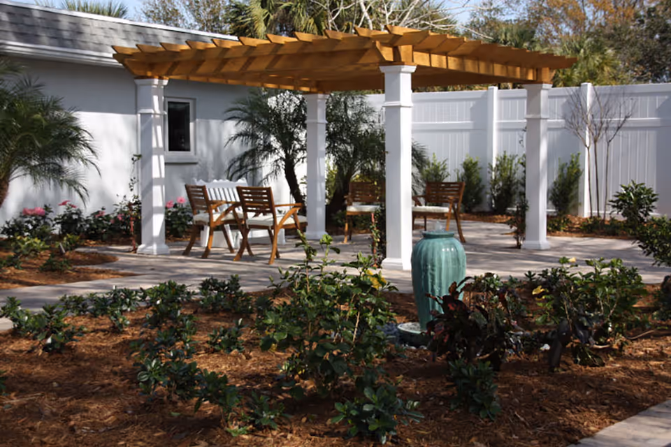 Landscaped outdoor patio featuring a wooden pergola with seating and a central green ceramic urn surrounded by plants.