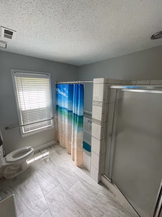 A bathroom with a toilet, a window with blinds, a shower with a sliding glass door, and a bathtub with a beach-themed shower curtain. The floor is tiled in a light color and the walls are painted gray.
