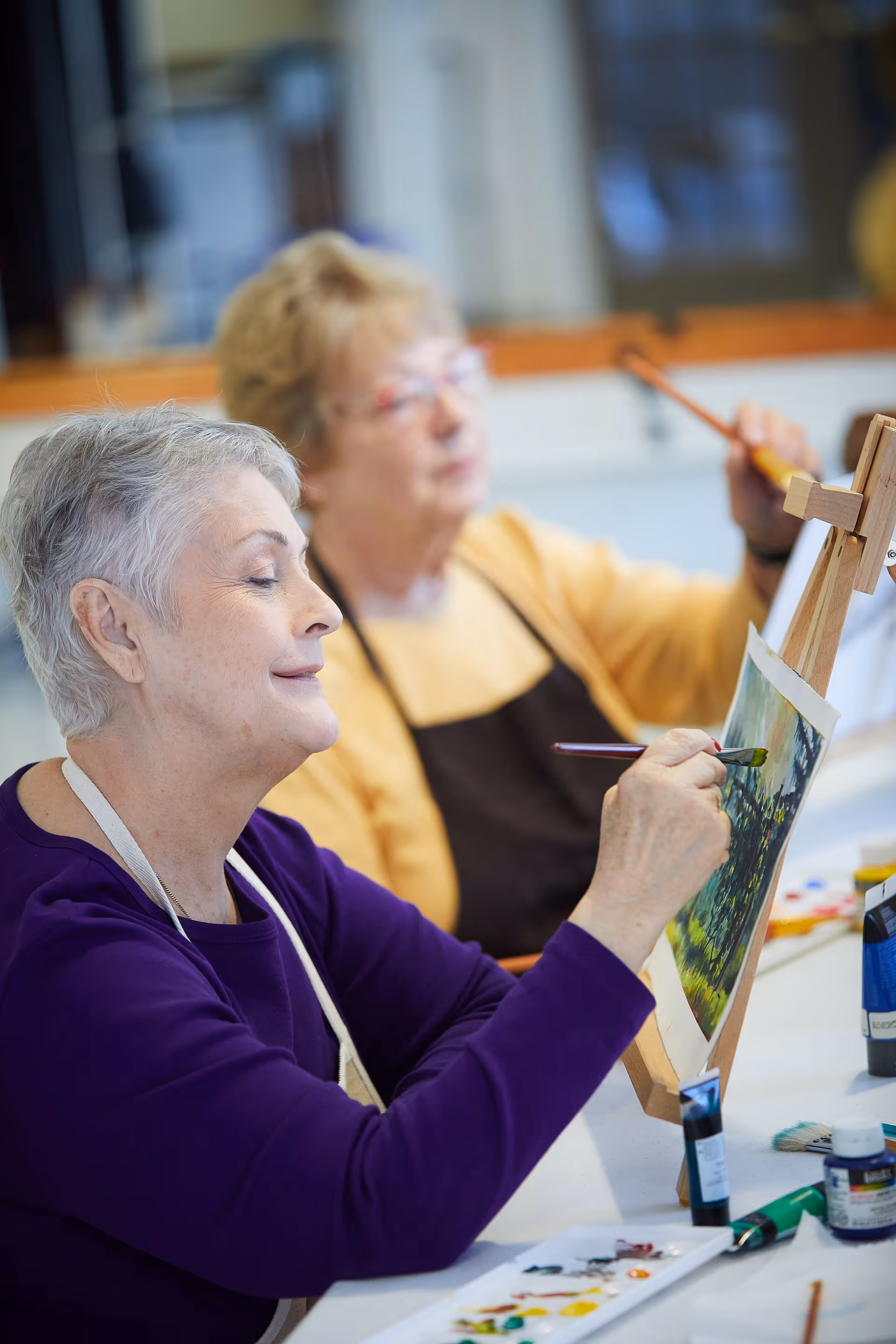 Two elderly women sitting at a table, painting on canvases placed on easels. One woman in the foreground is smiling and focused on her painting, while the other woman in the background is also painting. Various paint tubes and brushes are on the table.