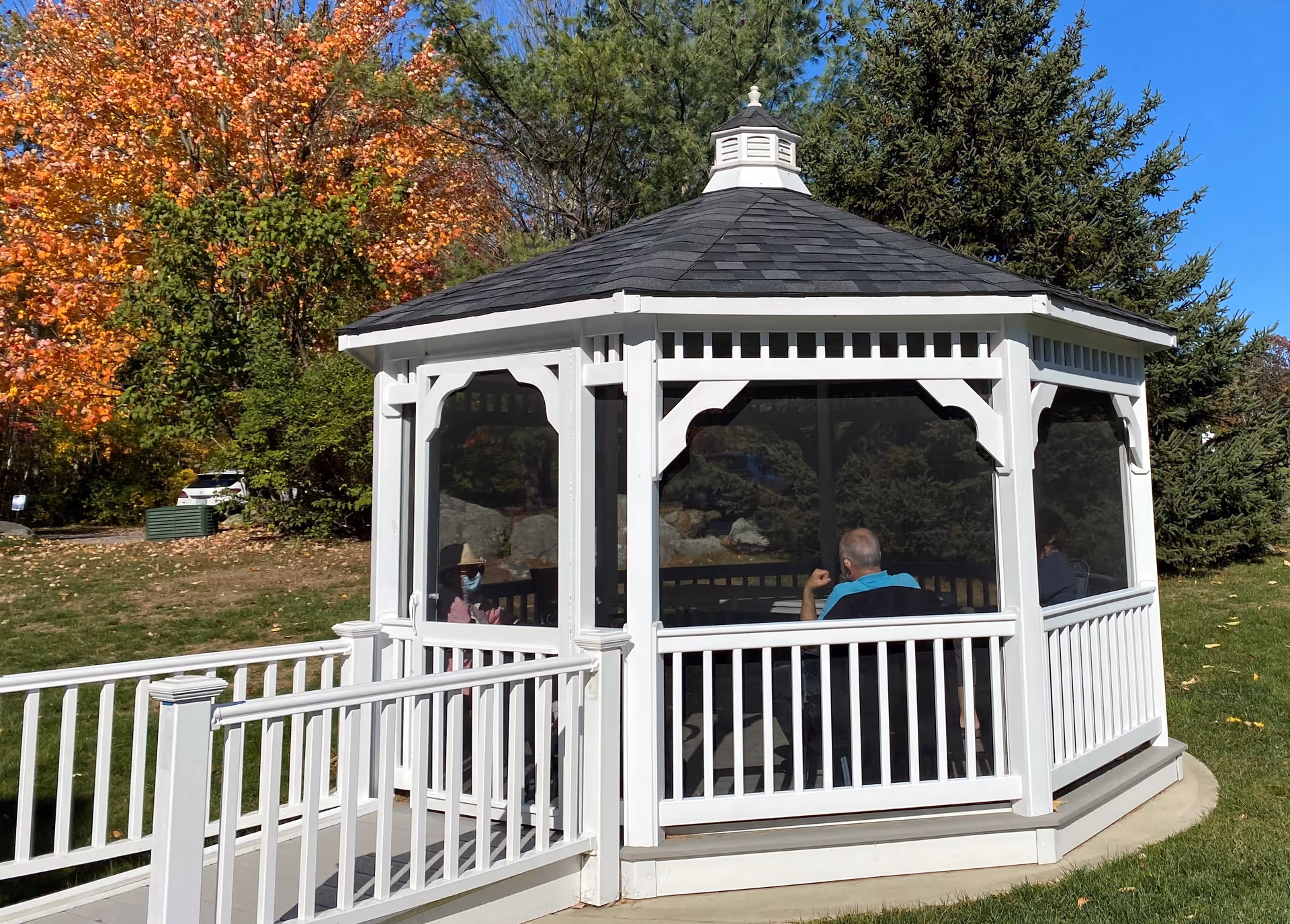 A white gazebo with a black shingled roof situated outdoors on a grassy area. Inside the gazebo, three people are seated and engaged in conversation. The surrounding area features trees with autumn-colored leaves and a clear blue sky.