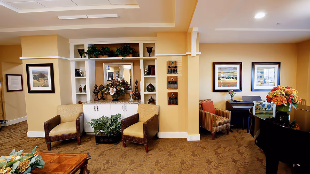 A cozy interior common area with beige walls and carpeted floor. The room features two cushioned armchairs with wooden arms, a built-in white shelving unit with decorative items and plants, a mirror, and a floral arrangement on a countertop. To the right, there is a striped armchair next to a piano with a vase of flowers on top. Framed landscape paintings hang on the walls.