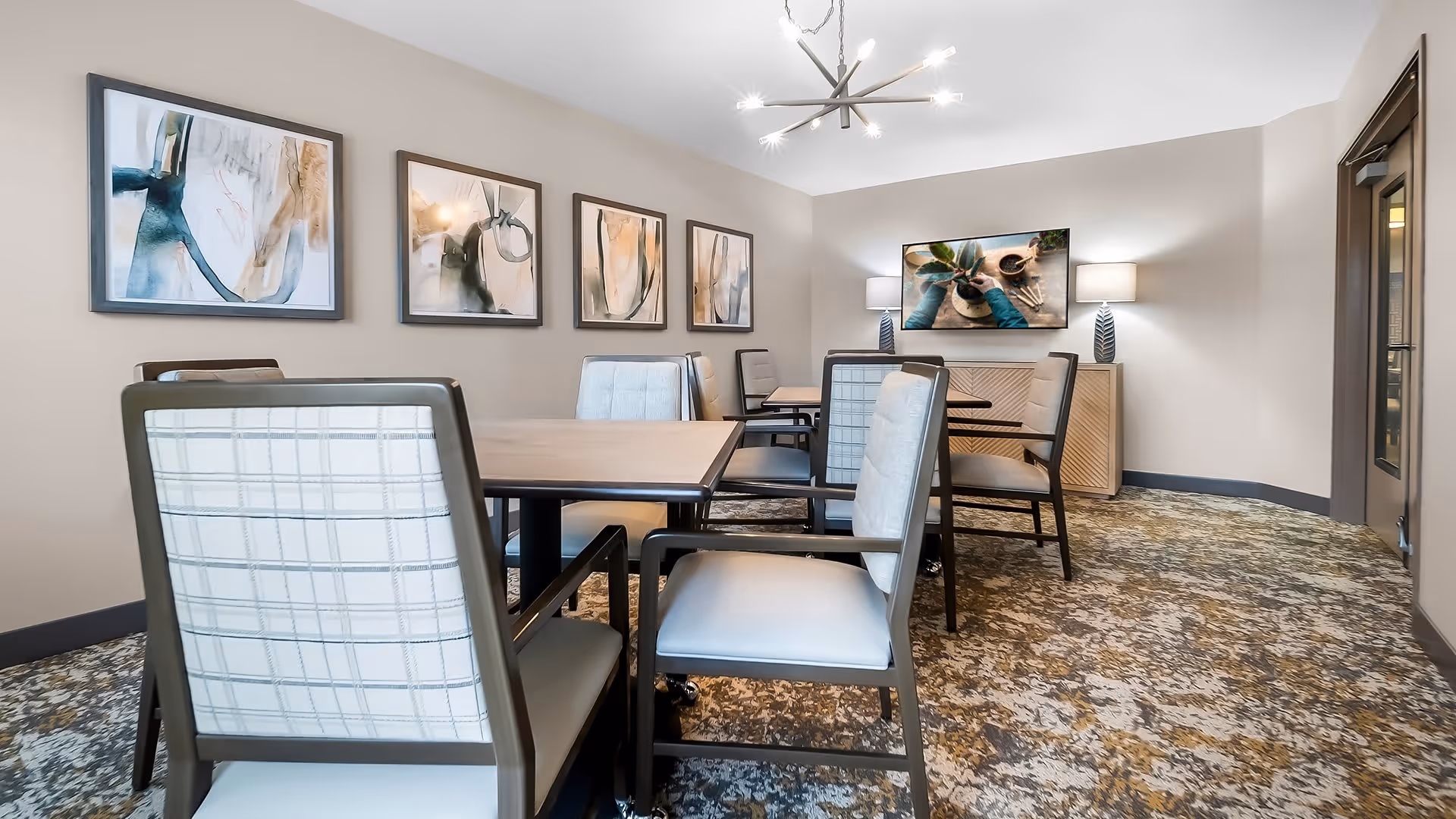 A modern dining room with several tables and cushioned chairs arranged neatly. The walls are decorated with abstract paintings, and there is a sideboard with two lamps and a framed picture above it. The room has a patterned carpet and a contemporary chandelier hanging from the ceiling.
