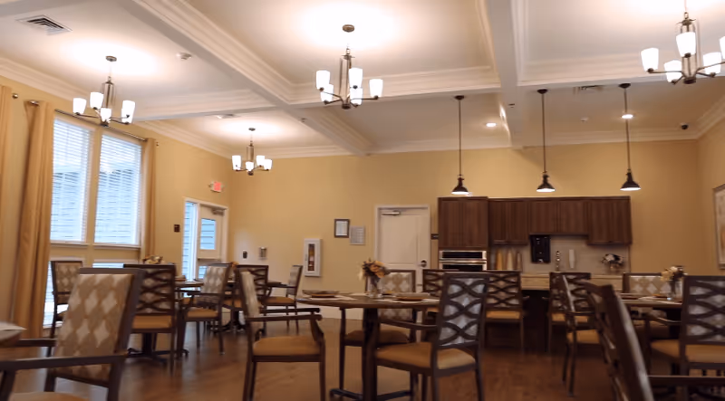A dining room in a senior living facility with multiple tables and chairs arranged neatly. The room features warm yellow walls, large windows with curtains, and ceiling lights including chandeliers and pendant lights. In the background, there is a kitchen area with wooden cabinets, a countertop, and built-in appliances.