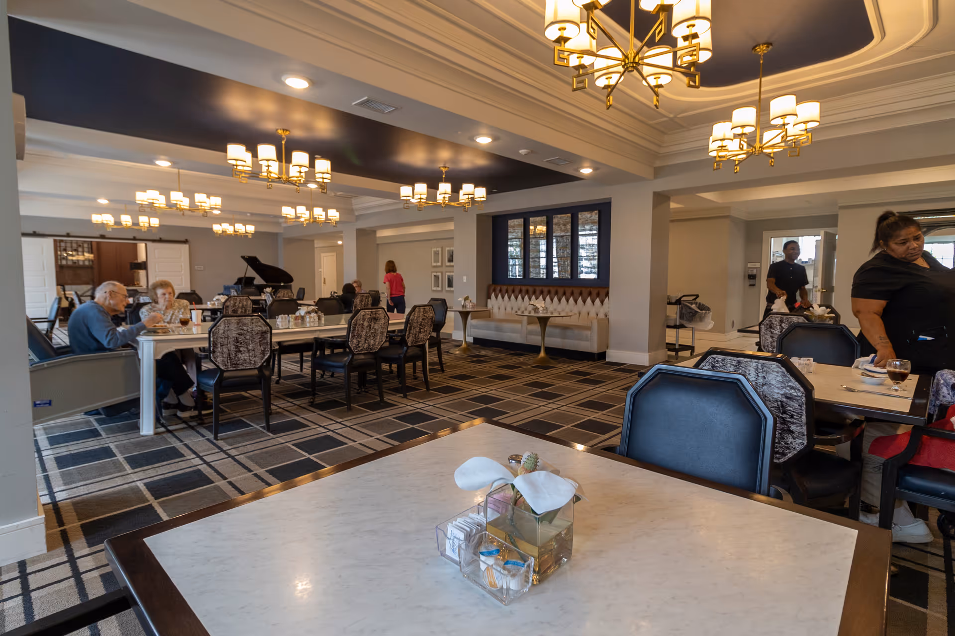 Spacious dining room with several tables and chandeliers, residents seated at a long table and staff moving about.