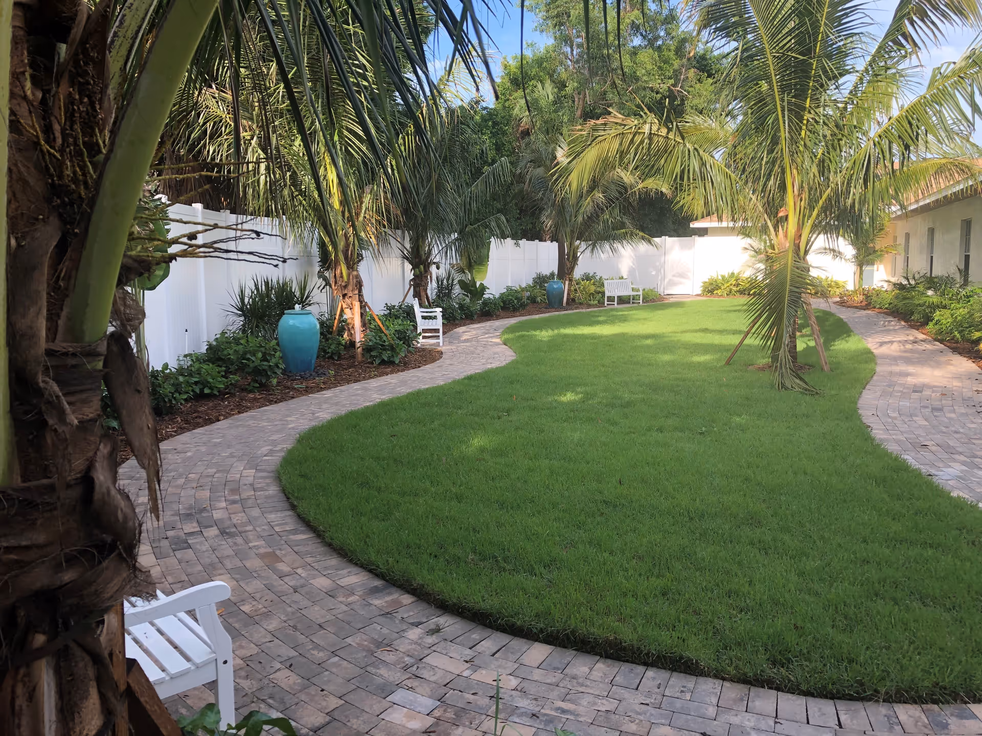 A serene outdoor garden area with a curved brick pathway surrounding a well-maintained grassy lawn. The garden is lined with palm trees, green shrubs, and large blue ceramic pots. White benches are placed along the pathway, and a white fence encloses the space.