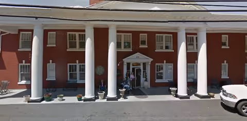 Front exterior view of a red brick building with white columns and a covered entrance. There are several windows with white shutters, potted plants along the front, and a few people near the entrance. A white vehicle is partially visible on the right side.