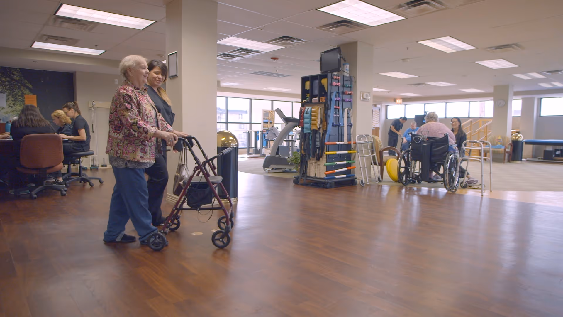 An elderly woman using a walker is assisted by a caregiver in a spacious room with wooden floors and large windows. In the background, other elderly individuals and caregivers are engaged in activities, including a person in a wheelchair and people seated at desks. The room contains exercise equipment and mobility aids.