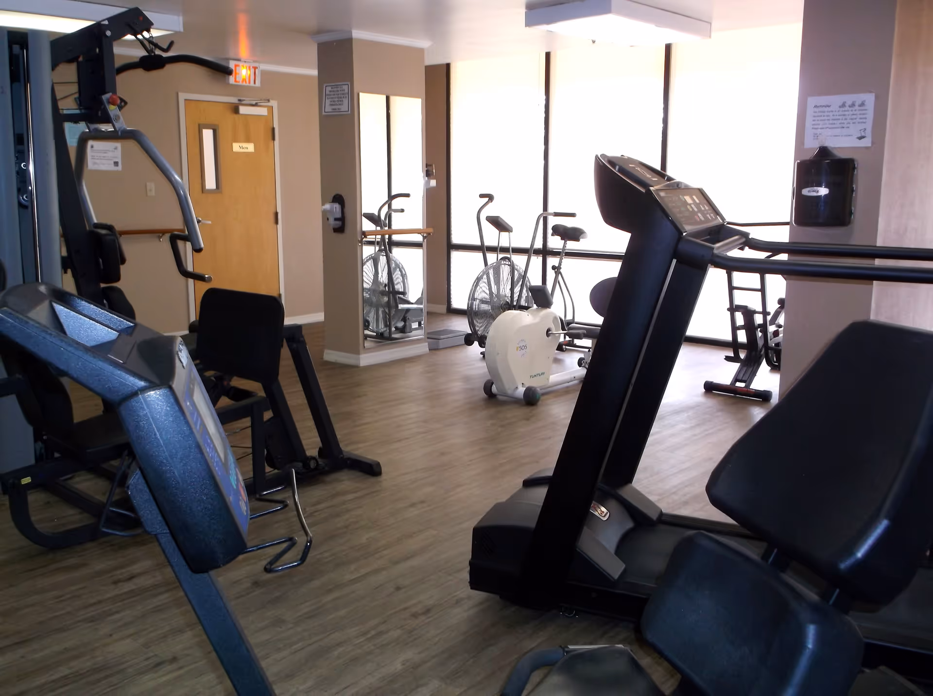 Interior view of a fitness room at Hampton Apartments with exercise equipment including a treadmill, stationary bikes, and weight machines. The room has wood flooring, large windows letting in natural light, a mirror on the wall, and a door labeled 'Men'.
