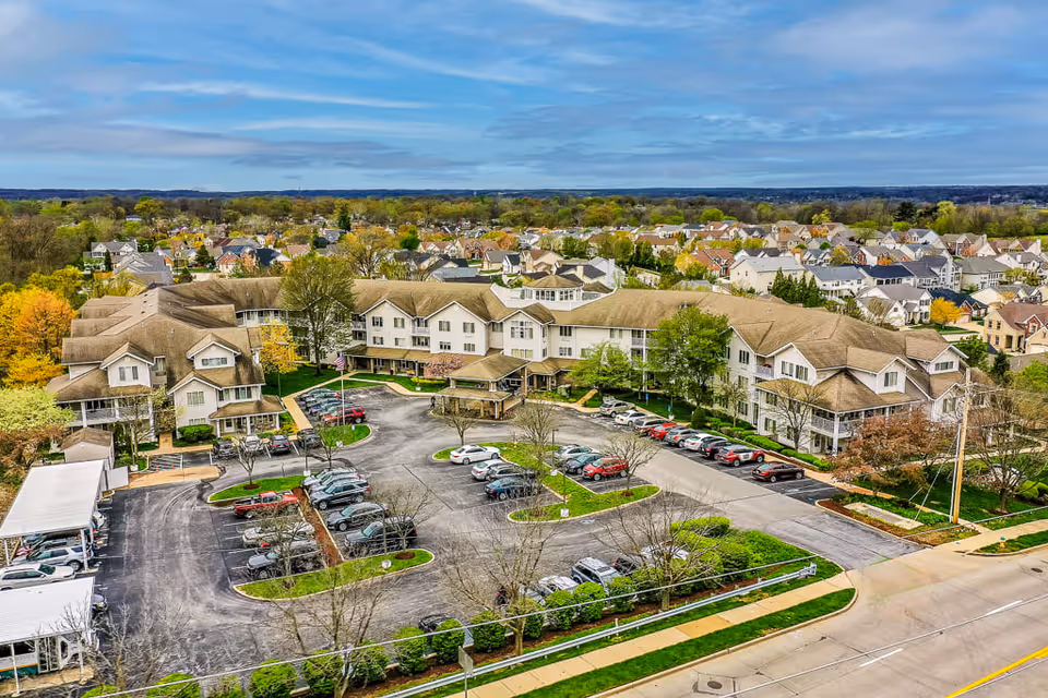 Aerial view of Solstice Senior Living at Fenton, showing a large multi-wing building surrounded by parking lots with numerous cars. The facility is located in a suburban neighborhood with many houses and trees visible in the background under a partly cloudy sky.