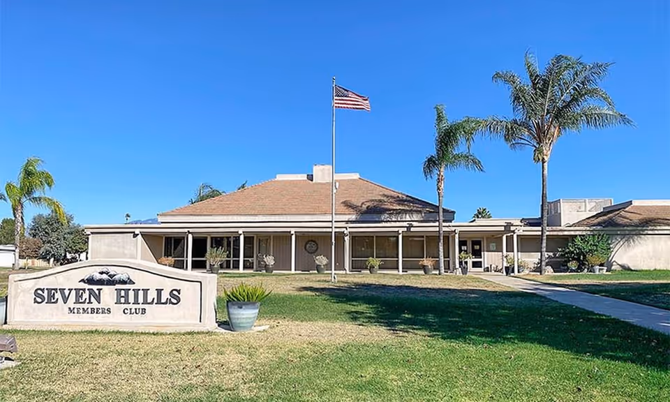 Front exterior of the Seven Hills Members Club building with a lawn, entrance sign, American flag, and palm trees.