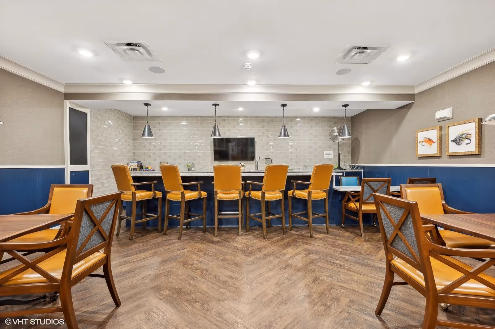 A modern dining area with a long counter featuring six mustard-yellow cushioned bar stools. Behind the counter is a wall with white subway tiles, a mounted flat-screen TV, and three pendant lights hanging from the ceiling. The room has wooden flooring and several wooden tables with matching chairs upholstered in mustard-yellow leather. The walls are painted beige with a blue wainscoting and decorated with framed artwork.