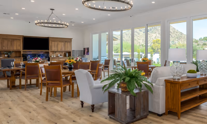A bright and spacious senior living common area with wooden tables and chairs arranged for dining, comfortable armchairs, and a sofa. Large windows offer a scenic view of mountains and greenery outside. The room features wooden flooring, modern light fixtures, and decorative plants on tables.