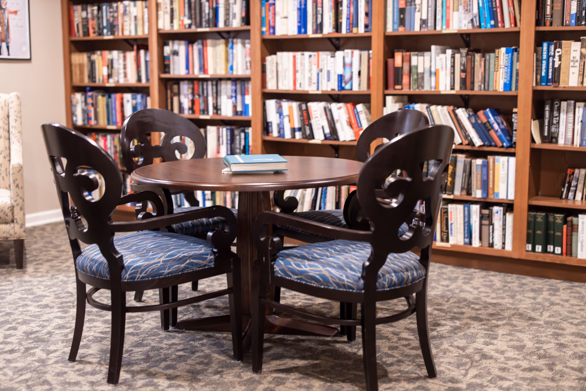 A cozy reading area in a senior living facility featuring a round wooden table with four dark wooden chairs upholstered in blue patterned fabric. The background shows tall wooden bookshelves filled with books, and a carpeted floor with a subtle pattern.