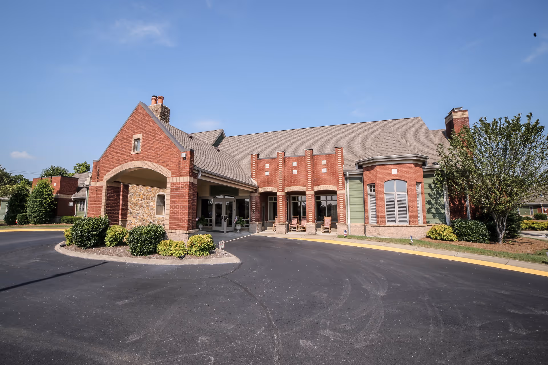 Front entrance of a brick senior living building with a covered porte-cochère, shrubs, and a driveway under a clear blue sky.