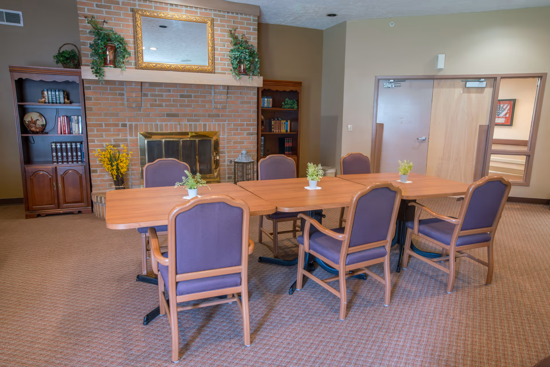 A meeting or dining room with a rectangular wooden table surrounded by six purple cushioned chairs. The room features a brick fireplace with a gold-framed mirror above it, flanked by two wooden bookshelves filled with books and decorative plants. The carpet is beige, and there are small potted plants on the table. A closed door and a window to another room are visible in the background.