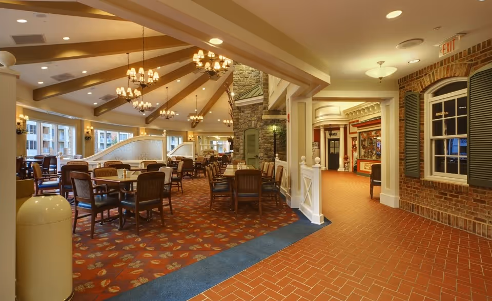 Interior view of a spacious dining area with multiple wooden tables and chairs arranged neatly. The ceiling features exposed wooden beams and several chandeliers with warm lighting. Large windows line the left side, allowing natural light to enter. To the right, there is a hallway with brick walls and decorative window shutters, leading to other parts of the facility.