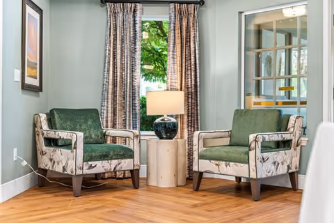 Two green cushioned armchairs with floral patterned sides are positioned on either side of a small round wooden table with a blue ceramic lamp on it. Behind the chairs is a window with patterned curtains, and the room has light green walls and wooden herringbone flooring.